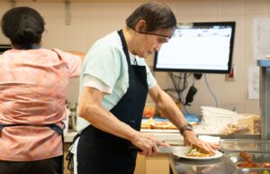 Two workers in a kitchen, in the foreground one is cutting a sandwich